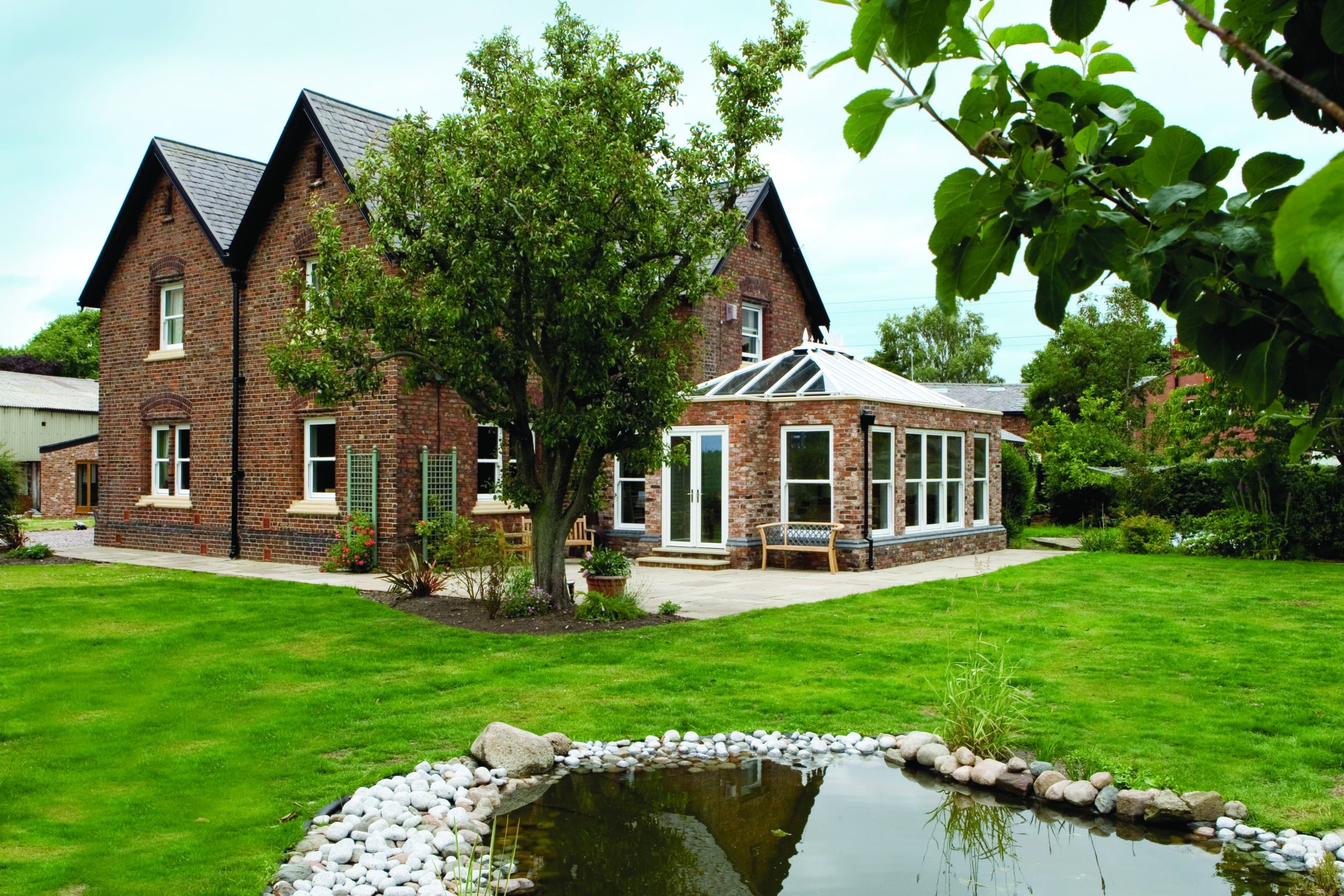 Orangery - A large brick house surrounded by trees, with a serene pond in the foreground.